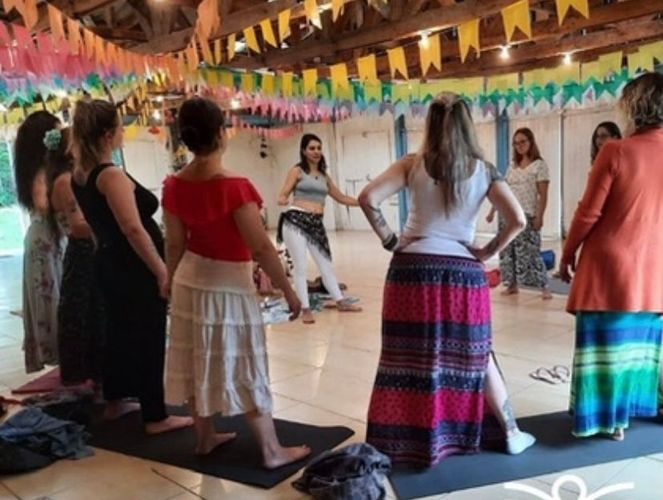 Mulheres em uma aula de dança, algumas observando uma dançarina, em uma sala decorada com bandeiras.