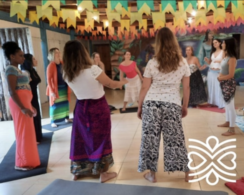 Grupo de mulheres em círculo, possivelmente em um retiro de ioga ou bem-estar, em um ambiente interno com decorações coloridas.