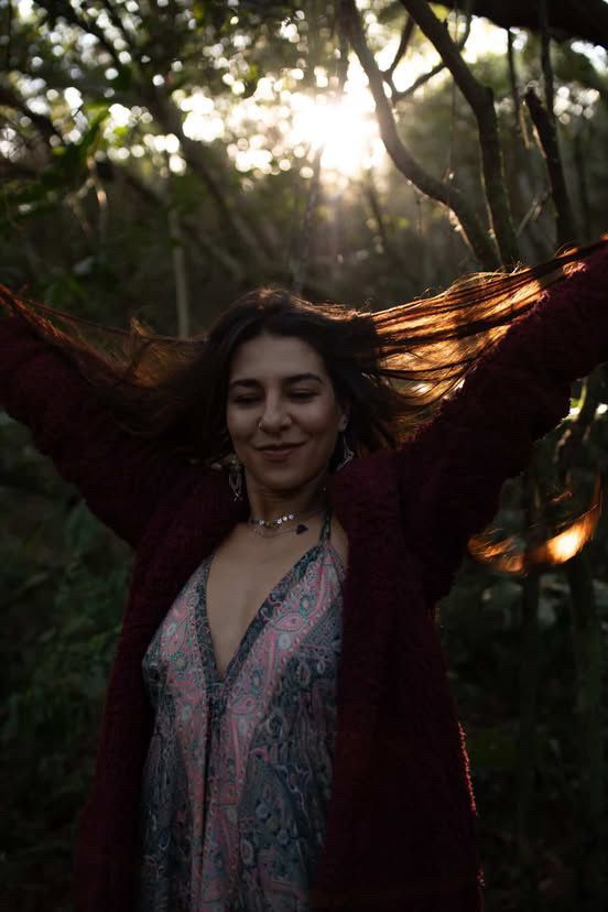 Mulher com os braços estendidos, cabelos castanhos balançando ao sol dentro de uma floresta.