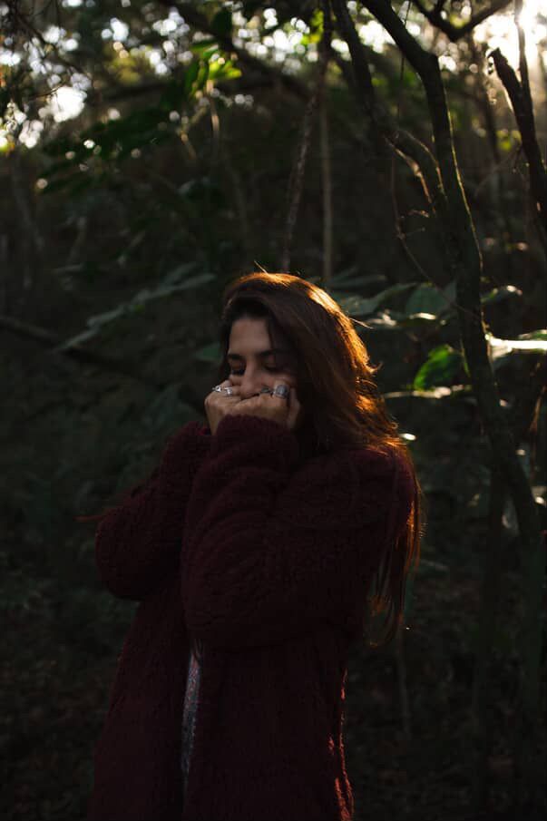 Mulher com longos cabelos castanhos, usando um suéter marrom, ao ar livre em uma floresta, com as mãos cobrindo a boca.