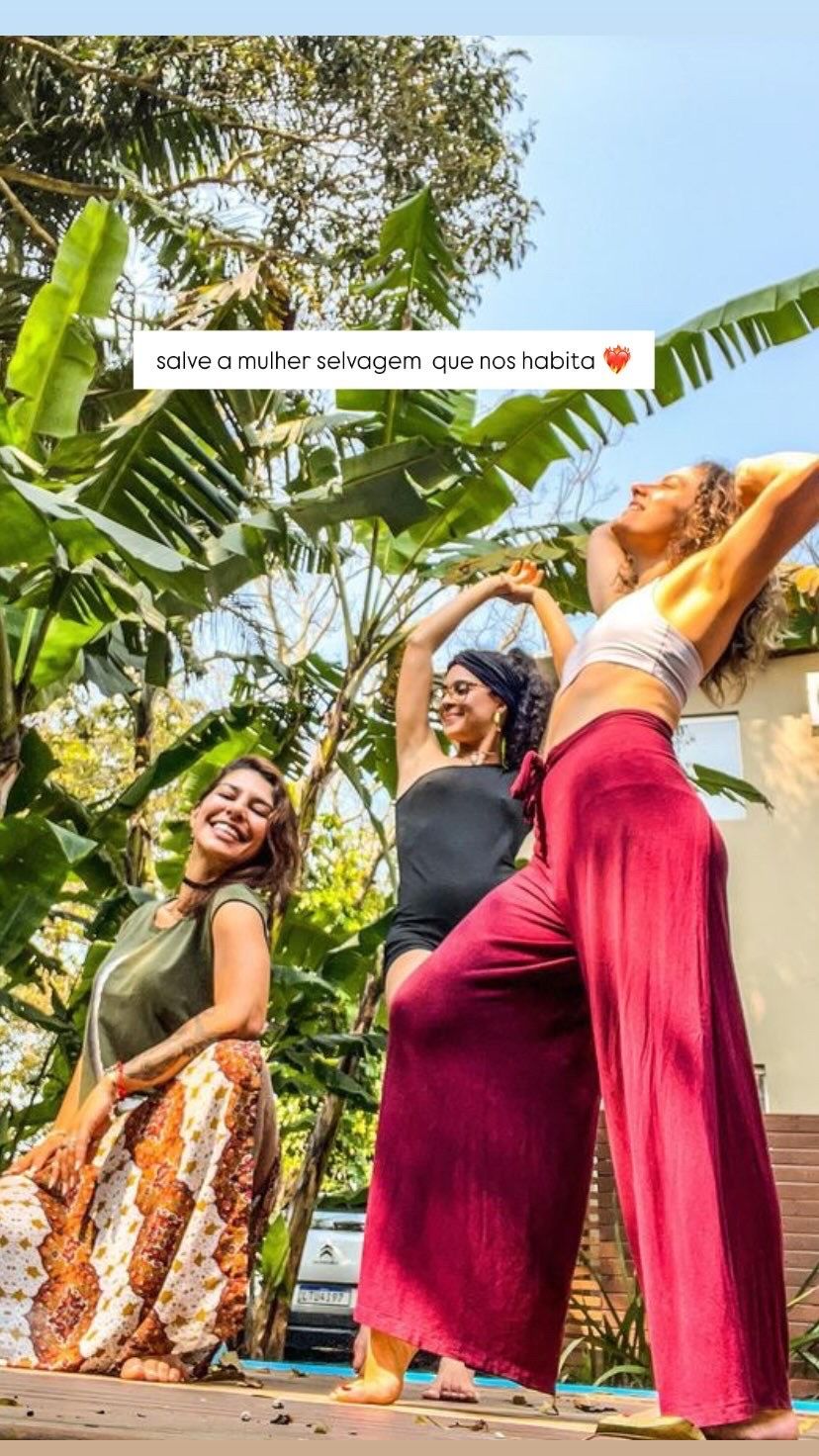 Três mulheres sorrindo e dançando ao ar livre com roupas coloridas; vegetação exuberante ao fundo.