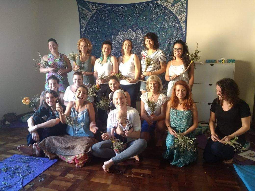 Grupo de mulheres segurando flores, sorrindo, em ambiente interno. Uma tapeçaria azul pendurada na parede.