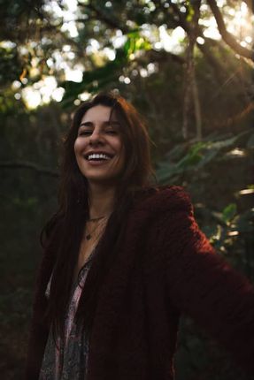Mulher sorrindo amplamente em uma floresta, vestindo um suéter vermelho, com a luz do sol filtrando através das árvores.