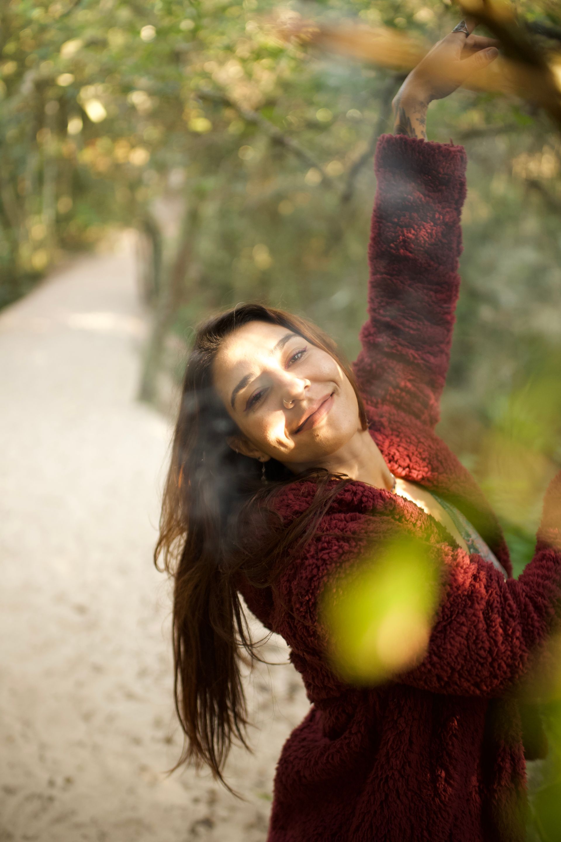 Mulher com suéter bordô sorri, alcançando um galho de árvore em um caminho arenoso através de uma floresta ensolarada.