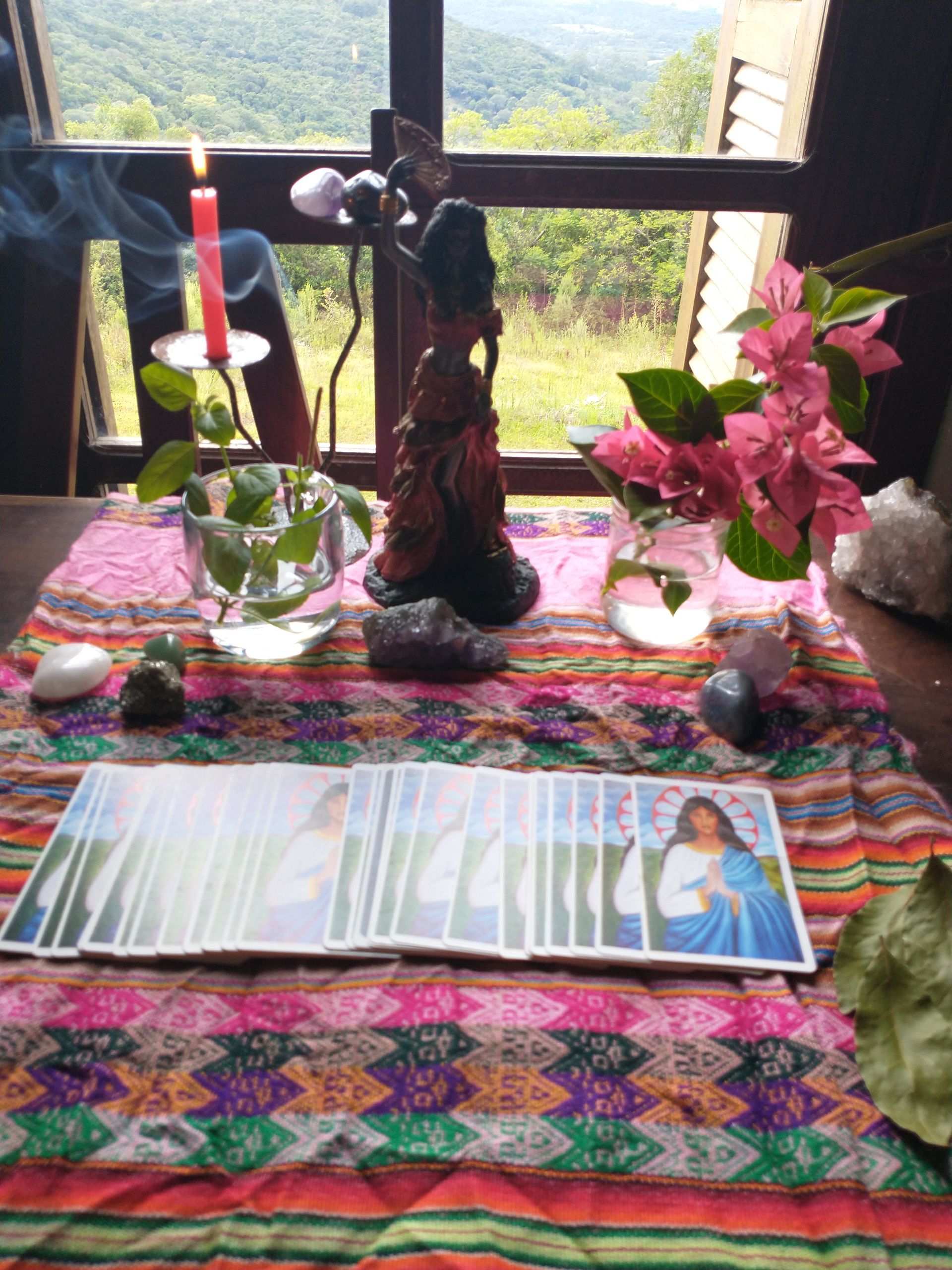 Altar com cartas de tarô, vela acesa, flores e estátua, colocado em frente a uma janela com vista para a montanha.