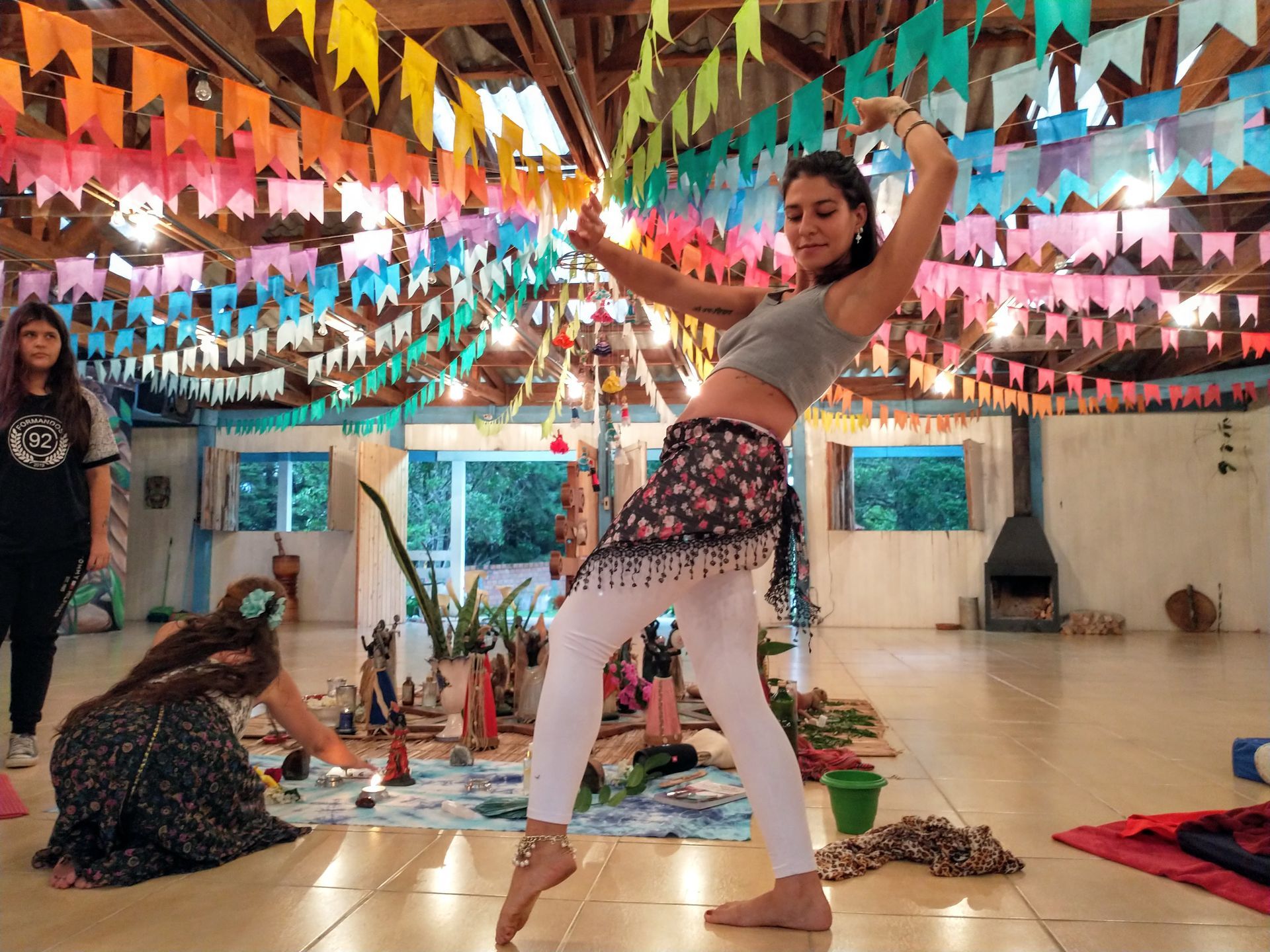 Mulher dançando com os braços levantados, em uma sala decorada com bandeiras coloridas; outras pessoas ao fundo.