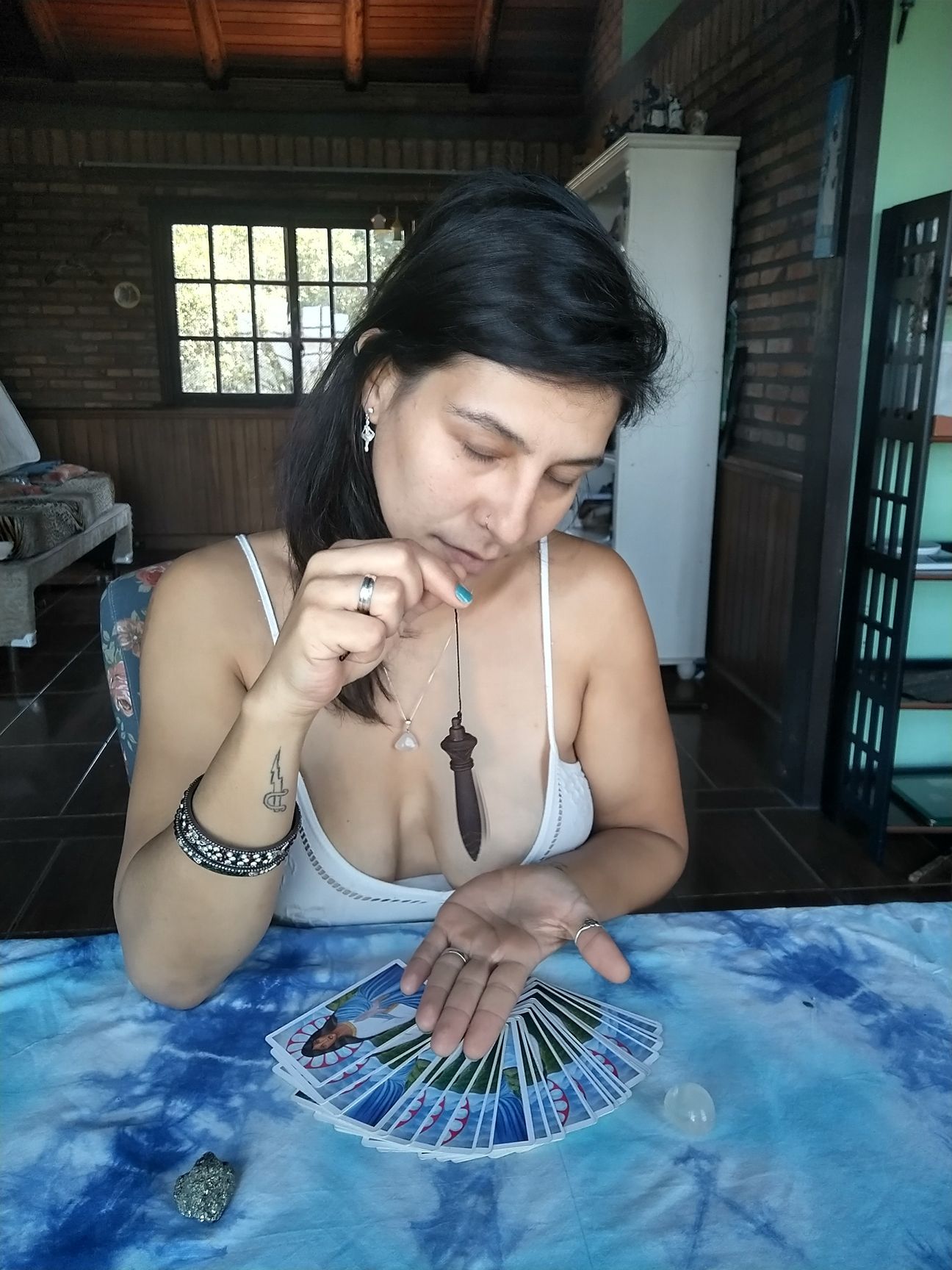 Mulher com cartas, pingente e cristal sobre uma mesa azul. Lá dentro, ela olha para baixo, pensativa.