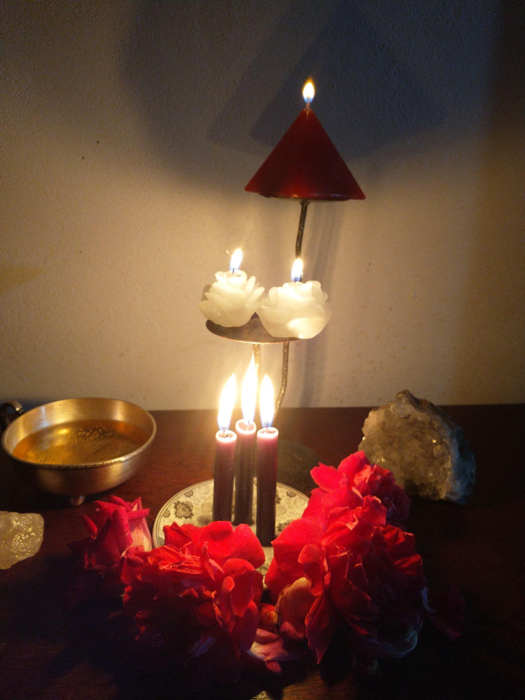 Velas e flores vermelhas sobre uma mesa. Líquido dourado em uma tigela, cristais e um castiçal em camadas.