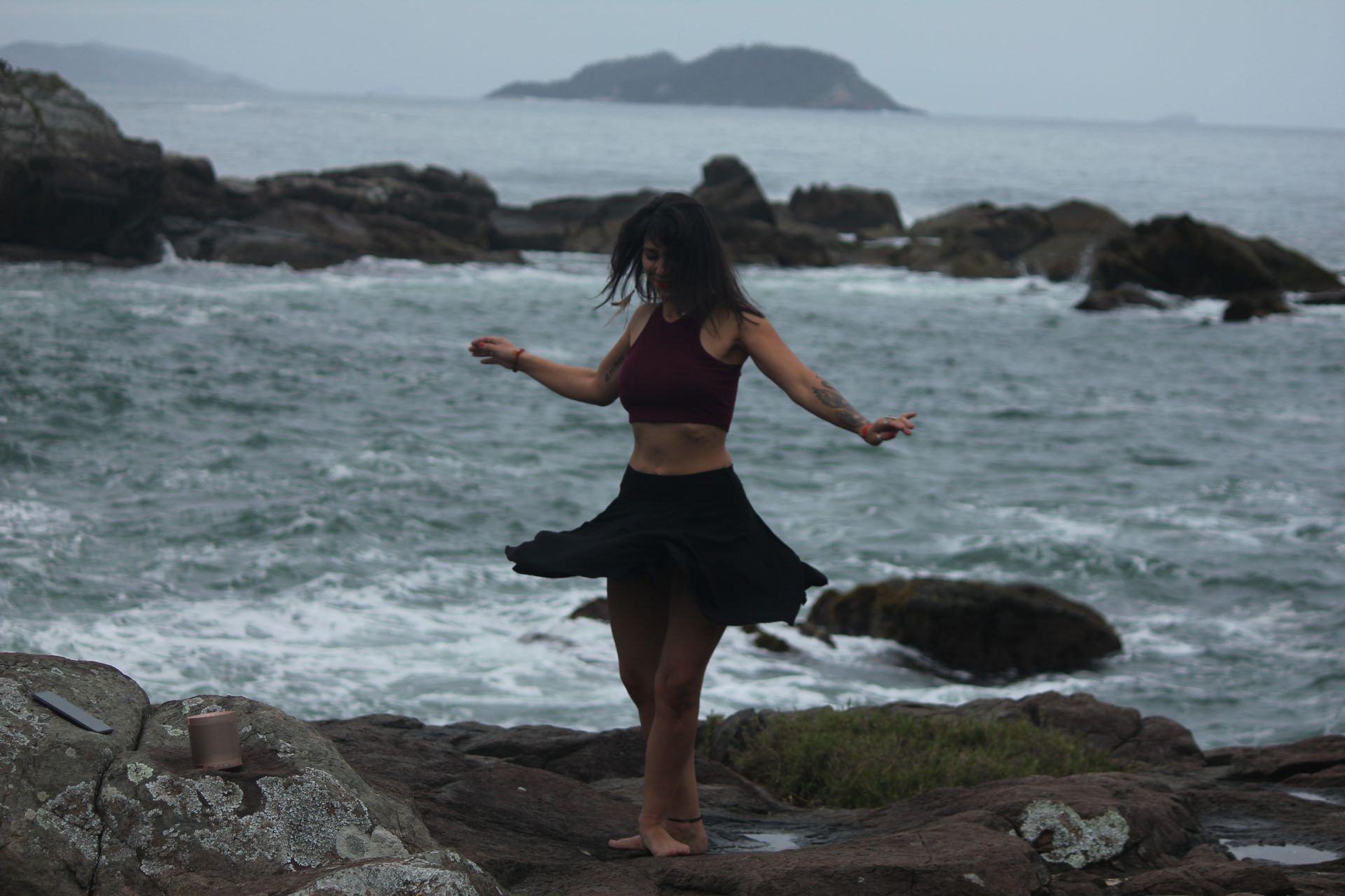 Mulher girando em uma praia rochosa, com o oceano ao fundo. Saia escura, blusa marrom, céu nublado.