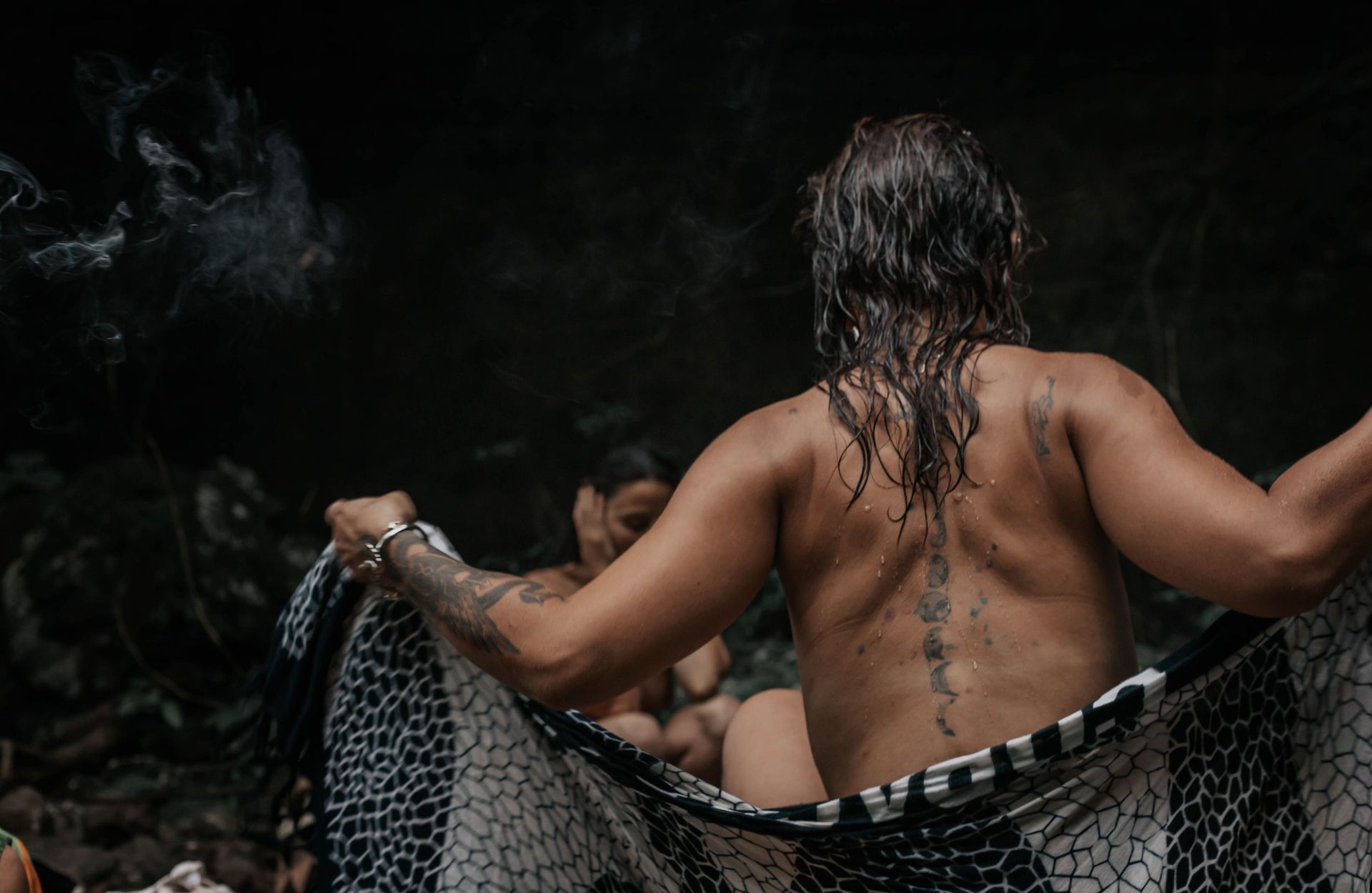 Homem com tatuagens cobre a si mesmo e a outra pessoa com um pano estampado perto de uma piscina escura e enfumaçada.