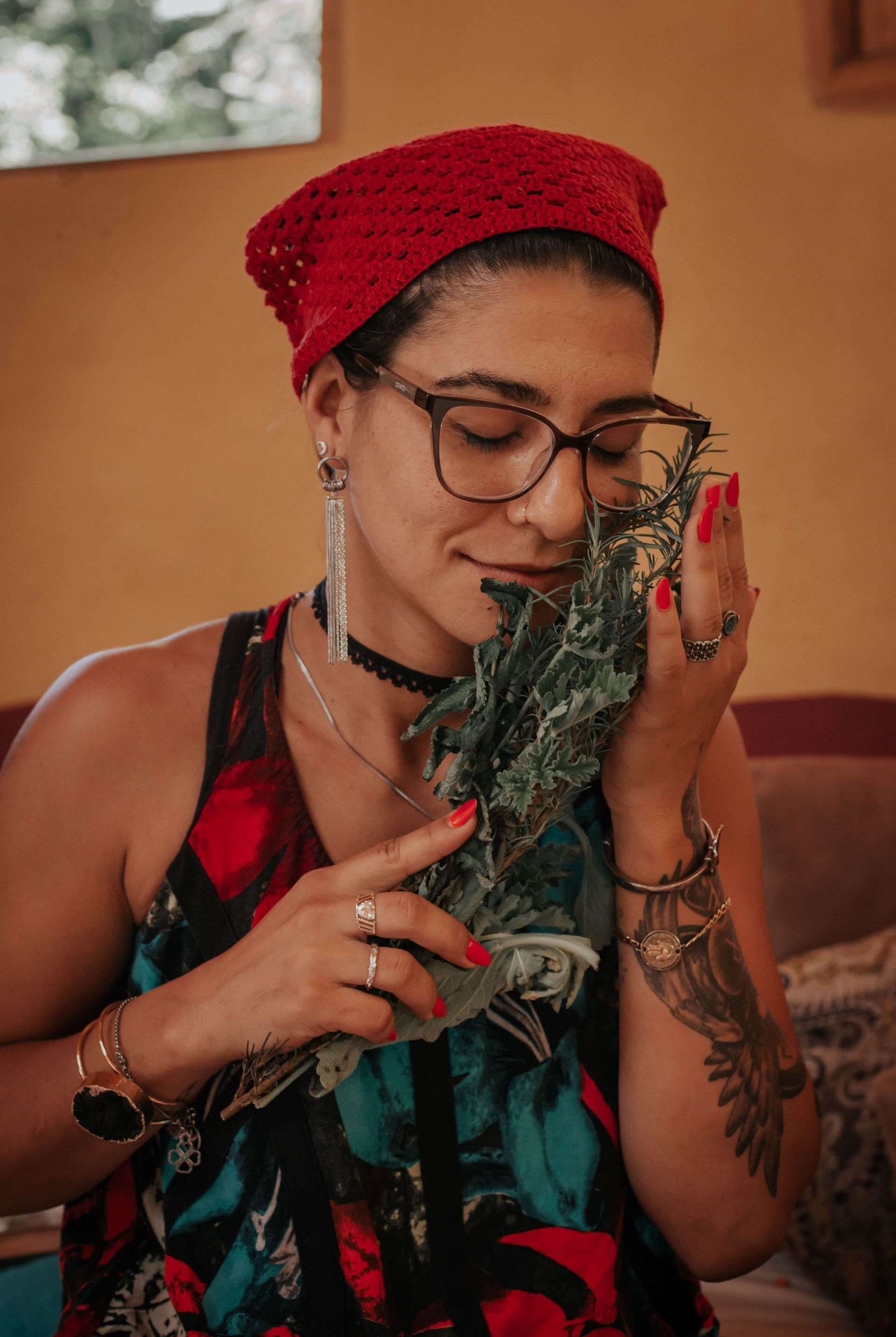 Mulher com lenço vermelho na cabeça cheirando ervas; usando óculos, joias e regata; dentro de casa.