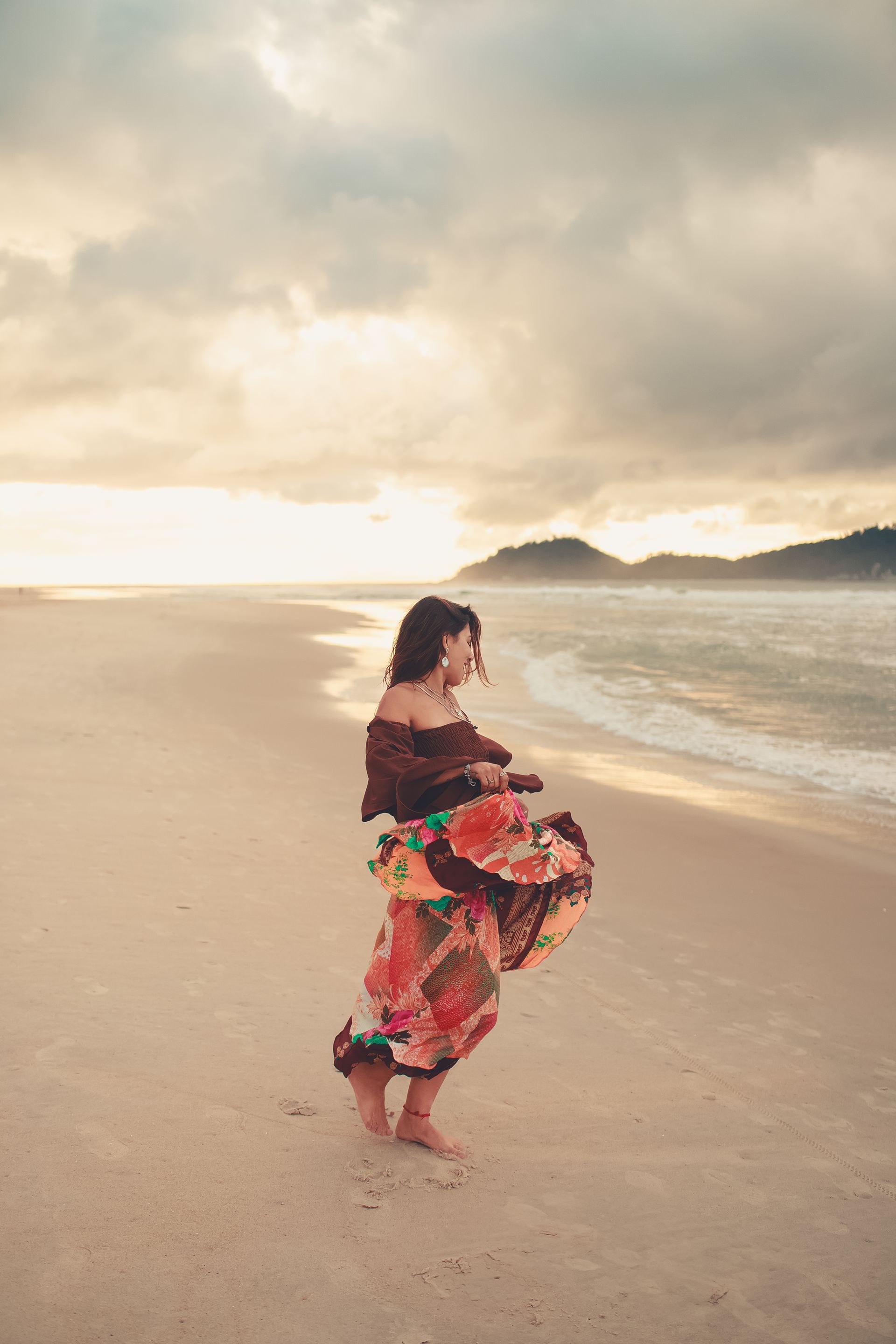 Mulher com vestido colorido girando na praia ao pôr do sol; ondas do mar, montanhas e céu nublado.