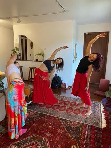 Três mulheres em uma sala se esticam de lado. Duas usam saia vermelha, uma saia estampada, e todas têm os braços acima da cabeça.