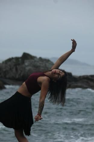 Mulher dançando em pedras perto do oceano, arqueando as costas, esticando os braços para cima, vestindo saia preta e blusa cor de vinho.