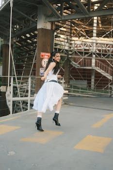 Mulher de vestido branco e botas pretas dançando em uma ponte, com estrutura de metal ao fundo.