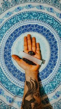 Mão segurando uma varinha de selenita, uma pedra escura e uma tatuagem, em um fundo de mandala azul.