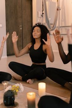 Grupo medita em um estúdio de ioga, braços erguidos. Mulher de roupa esportiva preta lidera, olhos fechados. Velas e flores no chão.