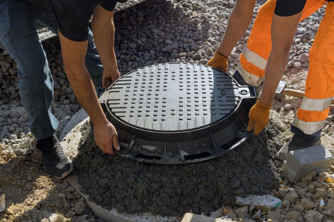 Two workers installing a manhole cover on a concrete base. One wears orange pants, the other blue jeans.