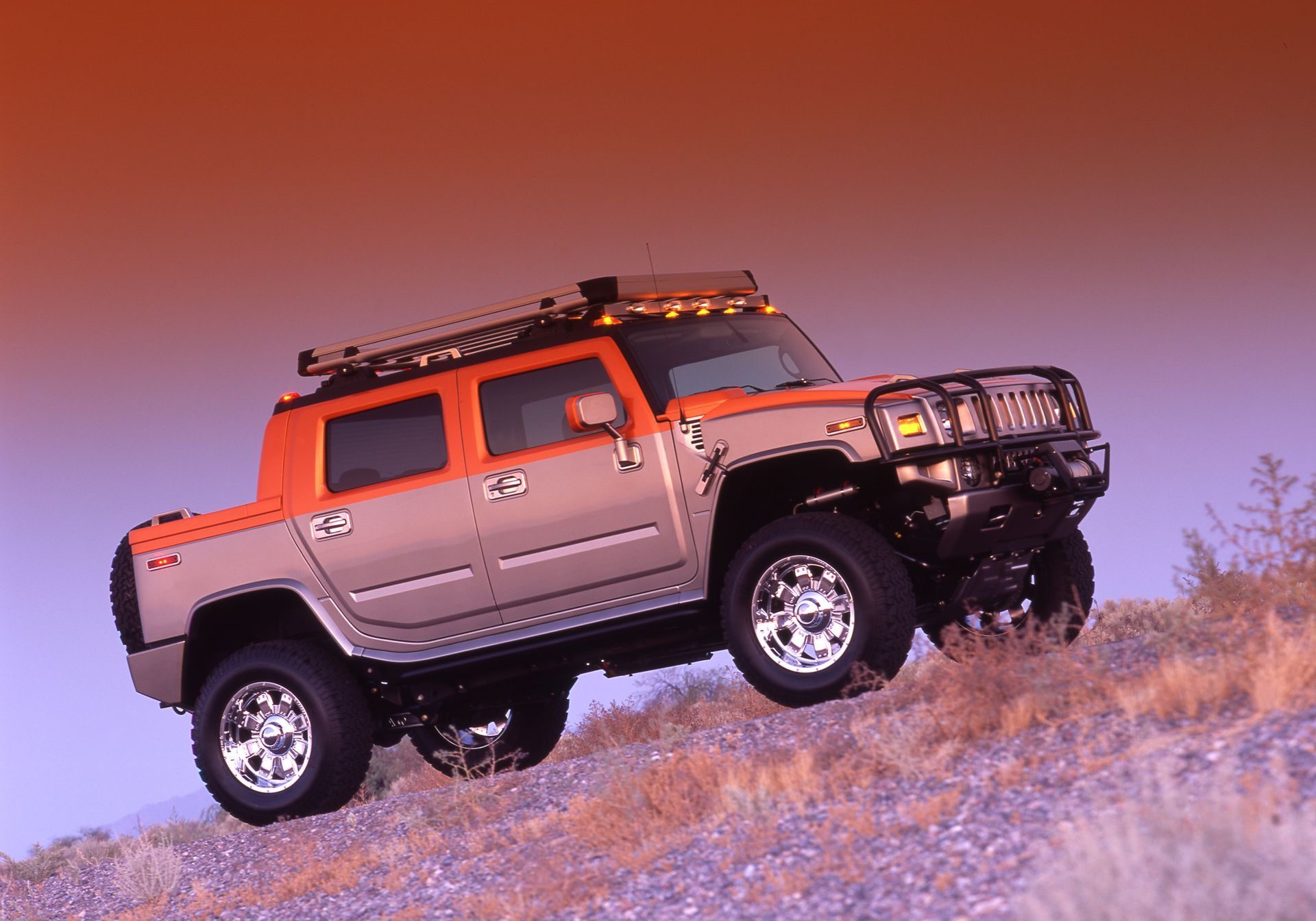 Orange and tan Hummer H2 truck driving uphill on a rocky, dry landscape at dusk.
