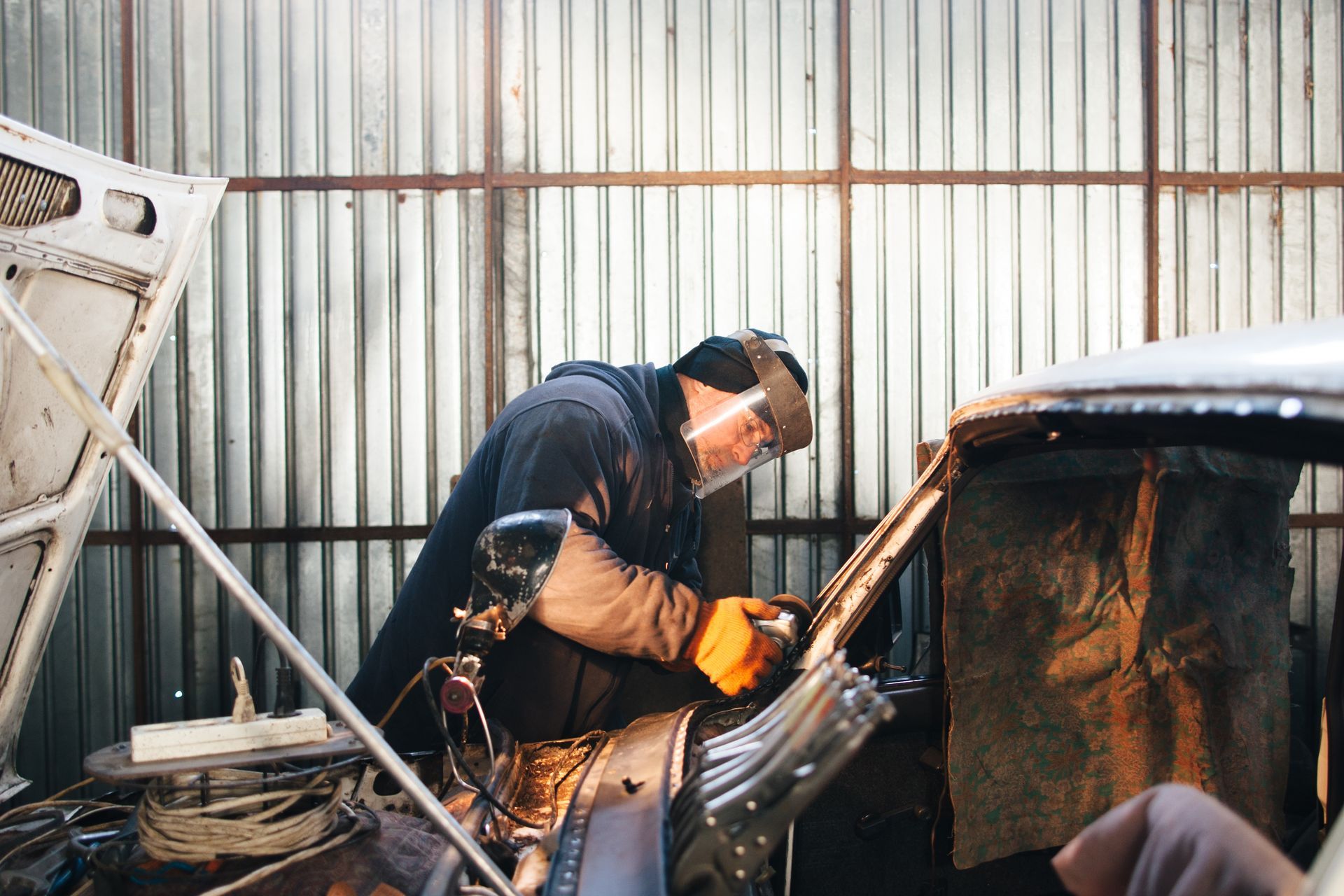 Person welds car in a garage, wearing protective gear, sparks visible.