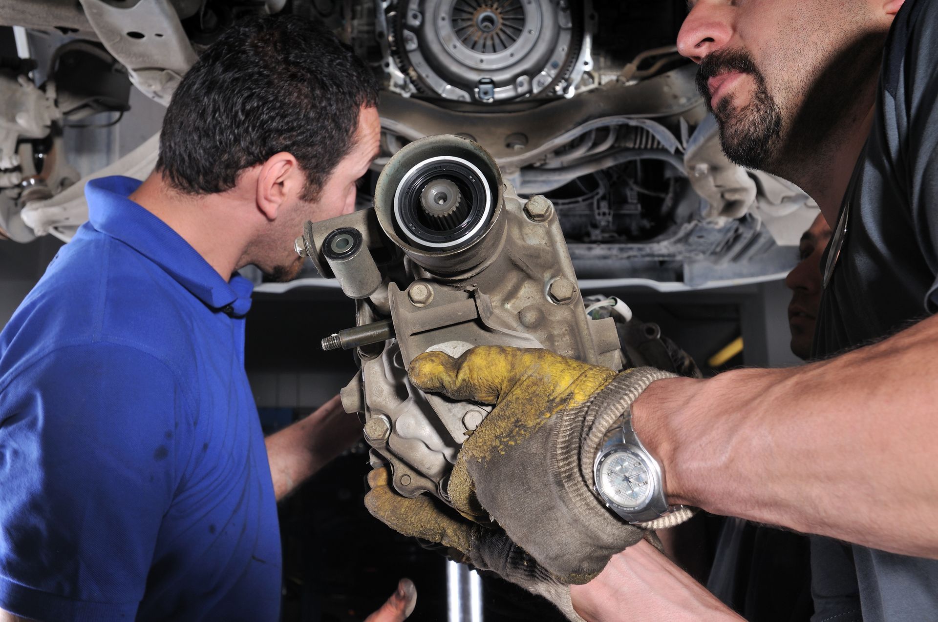 Two mechanics removing a car part. One wears a blue shirt, the other a glove. Working beneath the vehicle.