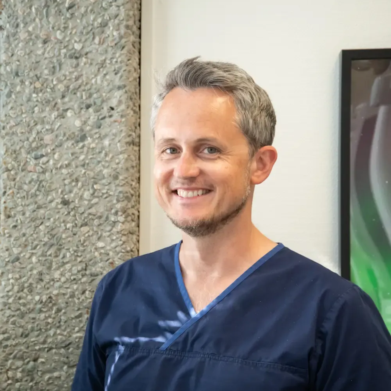 Tannlege Christian med briller og skjegg, iført blå skjorte og medisinsk maske, smiler til kameraet.