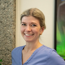 Kvinne med lyst hår smiler, iført blå skrubber, innendørs med nøytralfarget vegg og noe synlig dekor.