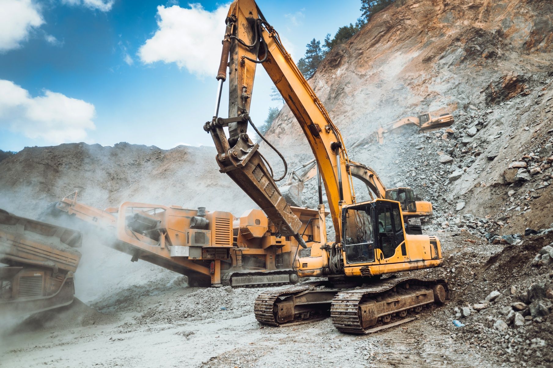 Yellow excavator breaking rocks at a quarry, with a backdrop of a rocky hillside under a blue sky.
