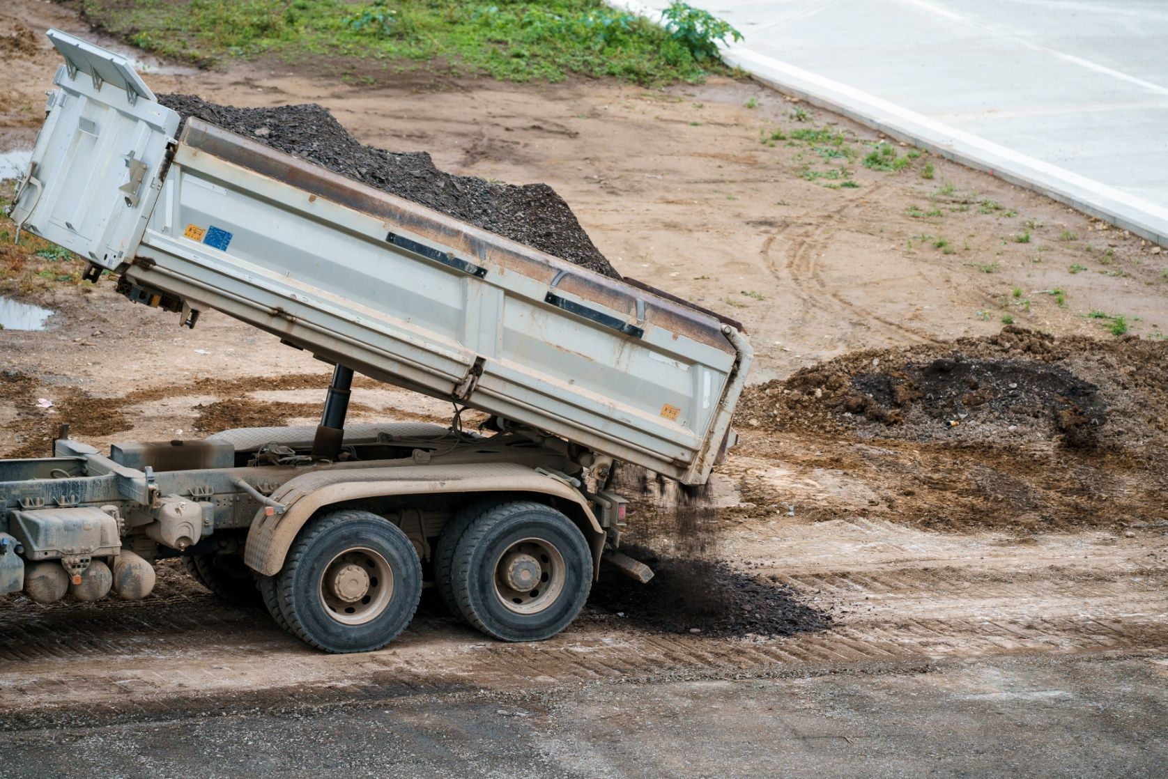Dump truck unloading dark material on a dirt ground next to a road.