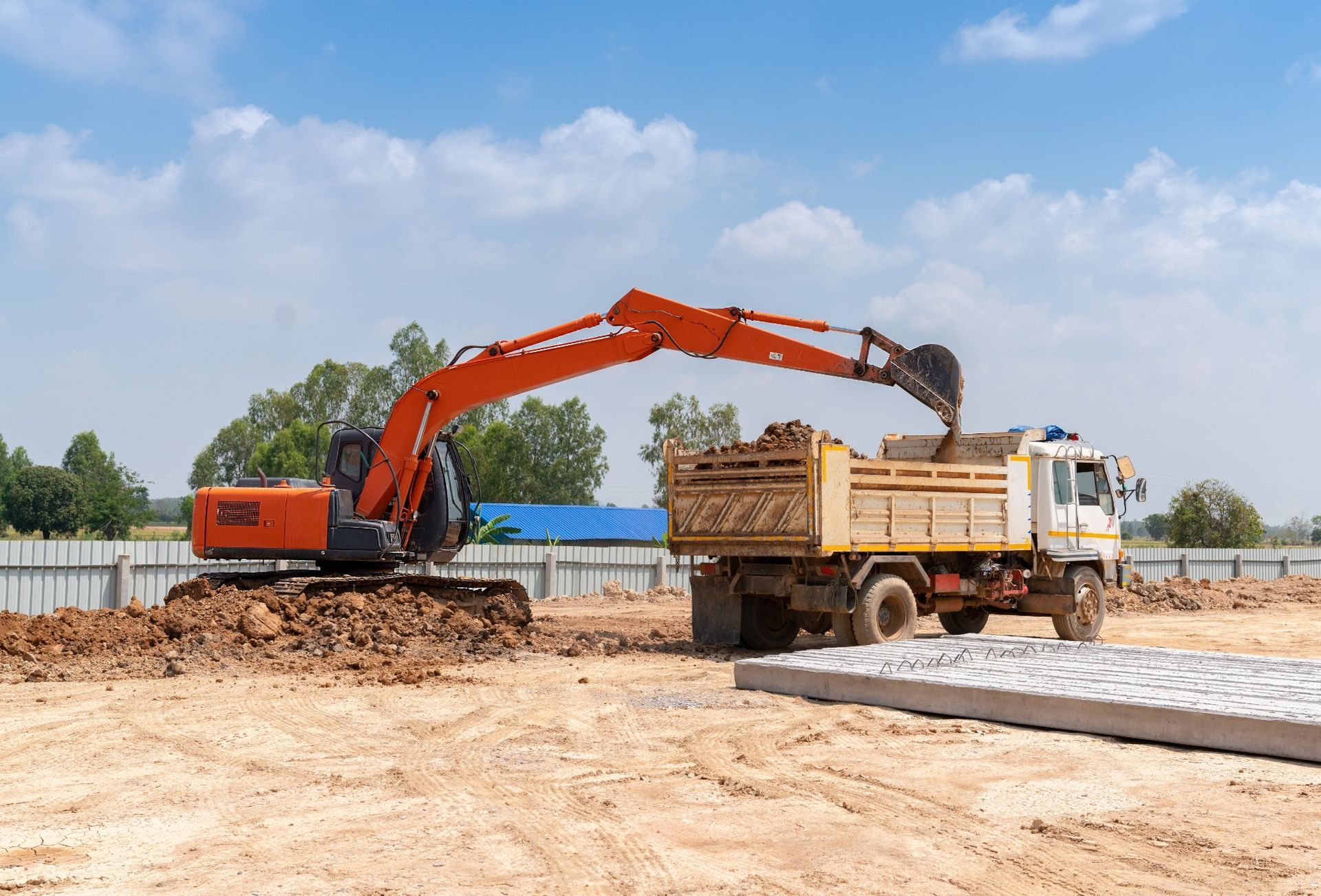 Orange excavator loading dirt into a white truck at a construction site.