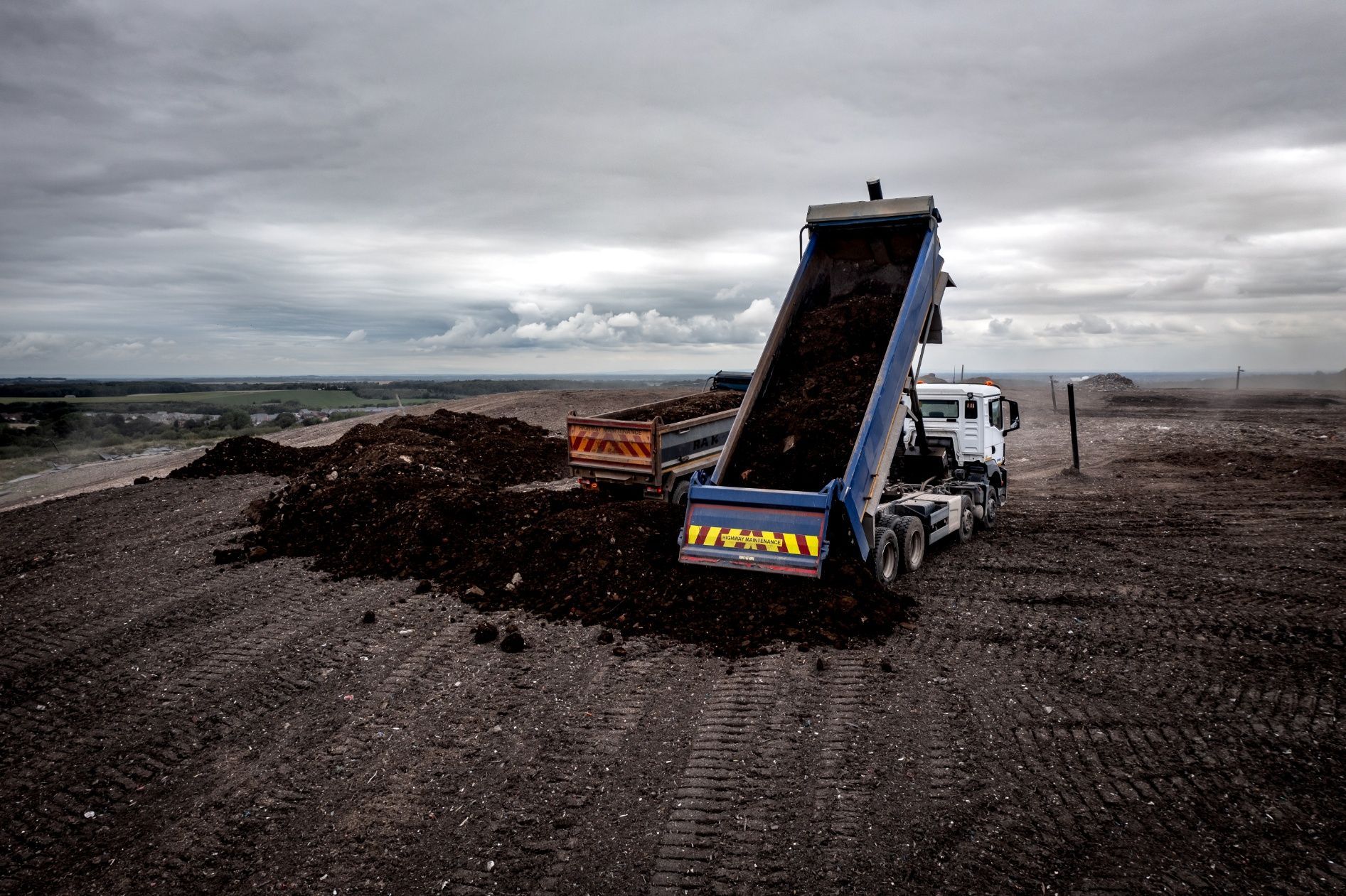 Dump truck unloading dark soil on a gray, overcast day at a landfill site.
