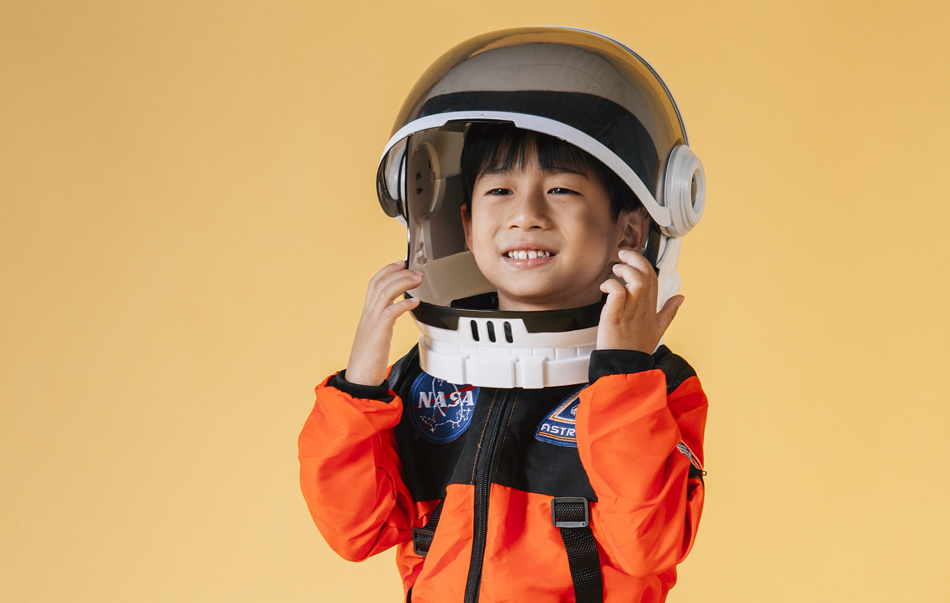 A young boy dressed as an astronaut is putting on a helmet.