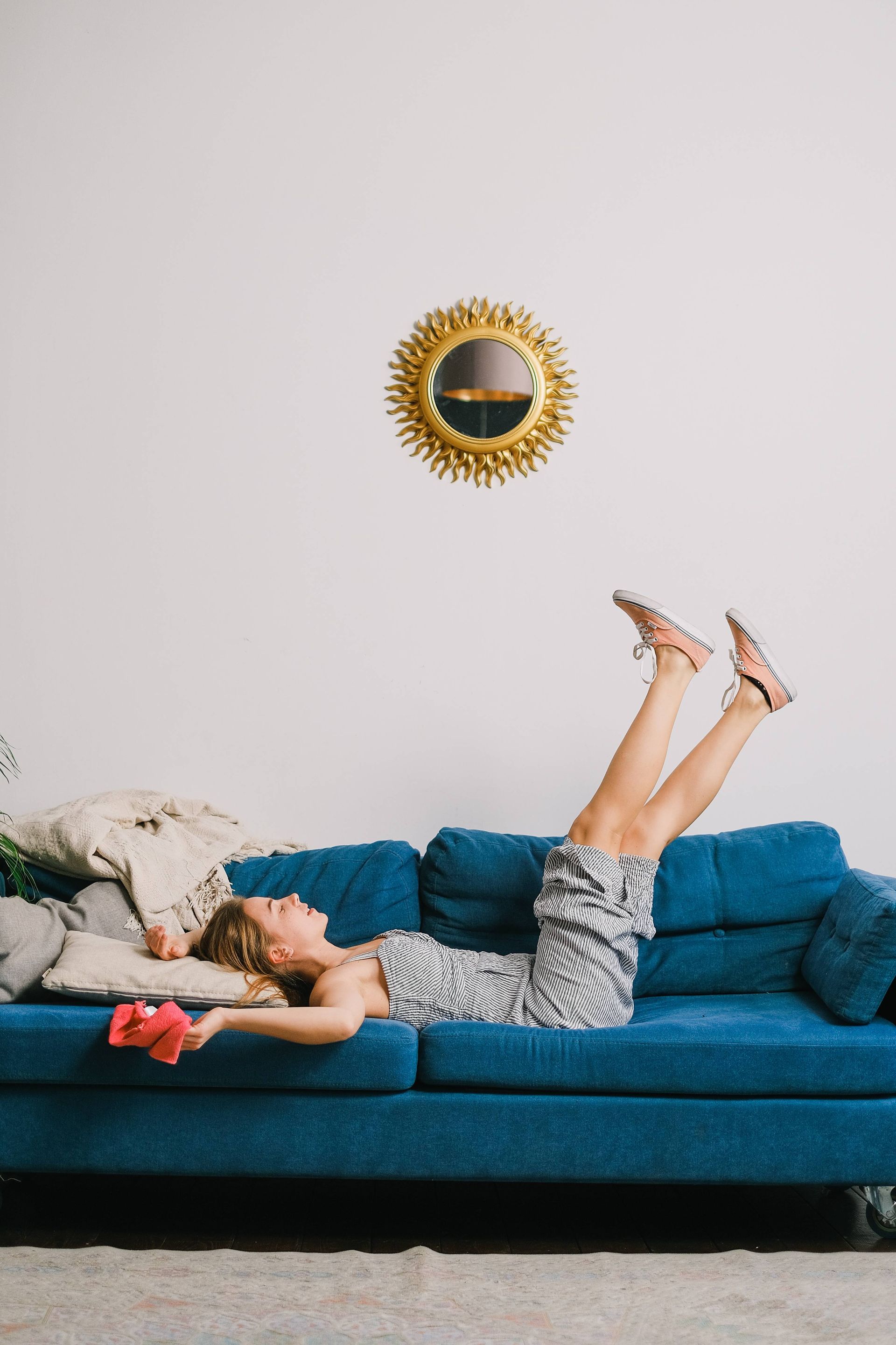 Woman lying on blue couch with legs up, holding a pink cloth, looking relaxed. Gold sunburst mirror on wall.