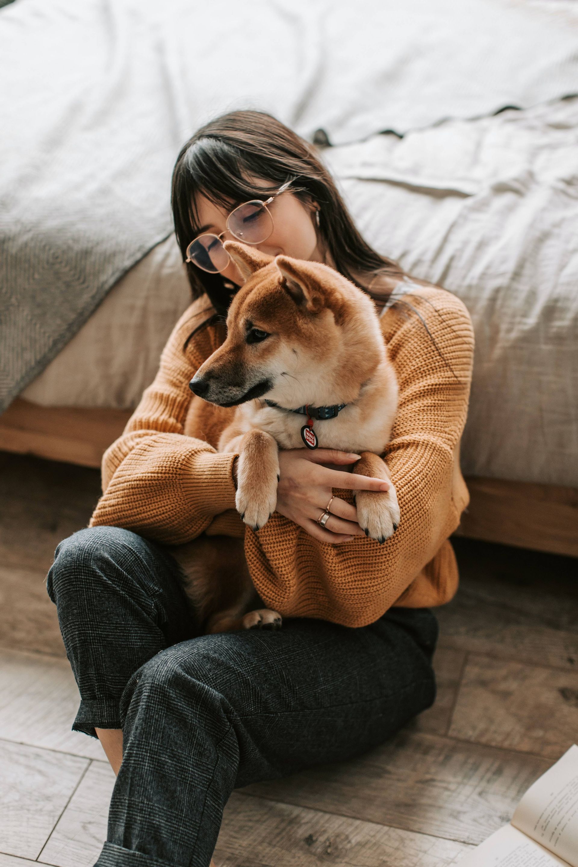 Woman in orange sweater hugs Shiba Inu dog on floor near bed.