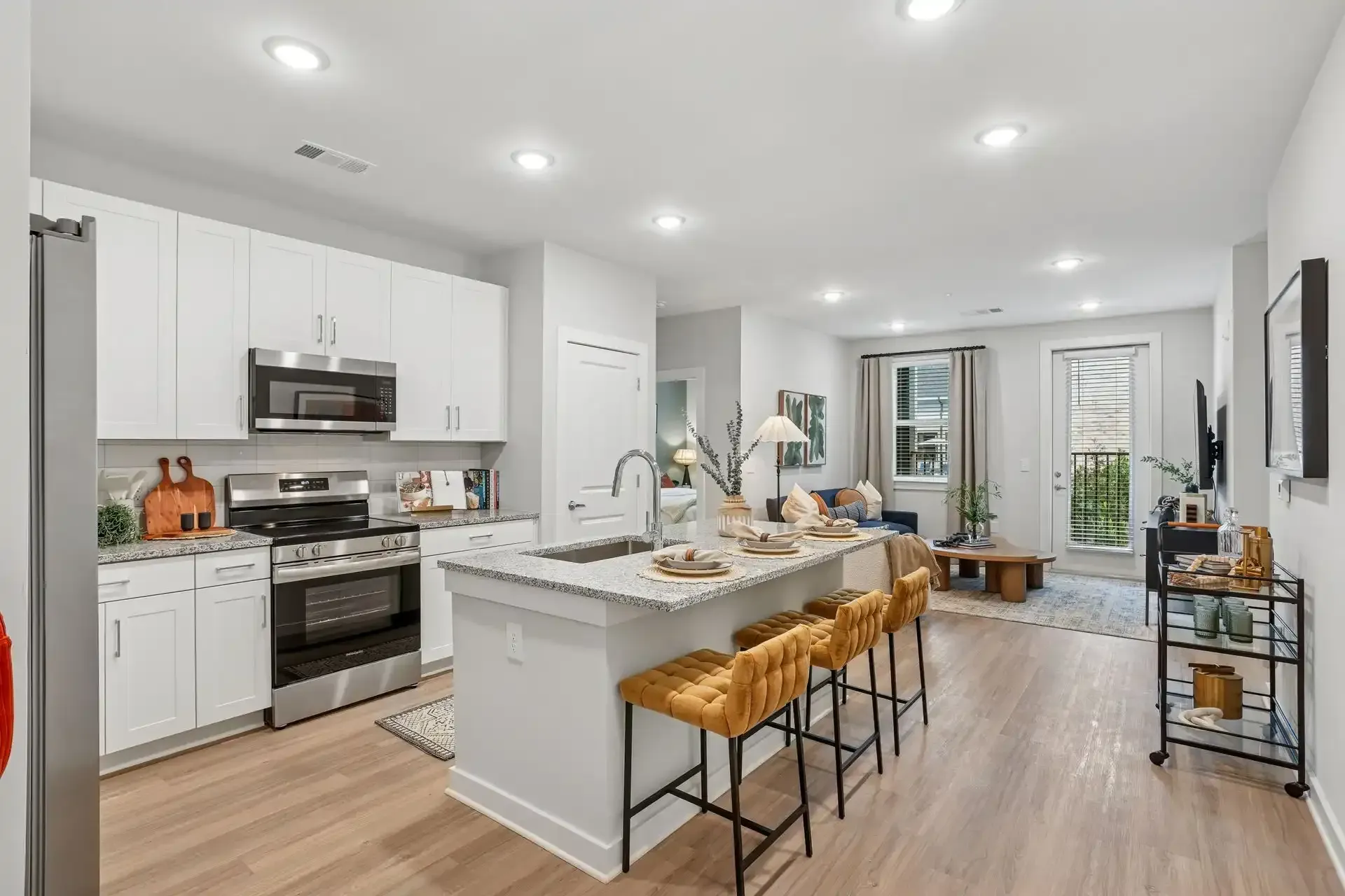 A modern open-concept kitchen with white cabinets, a granite island with four mustard-yellow bar stools, and hardwood.