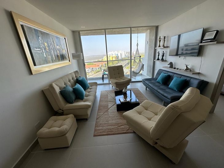 Sala de estar con muebles beige, cojines azules y vista a la ciudad. | Mevak Agency