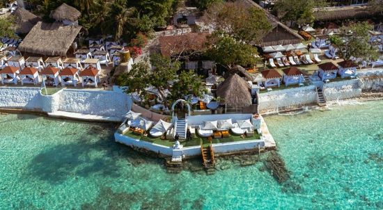 Vista aérea de un resort frente a la playa con cabañas blancas, agua turquesa y techos de paja. | Mevak Agency