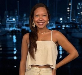 Mujer sonriente, vestida con un top color crema y joyas, frente a un puerto deportivo nocturno. | Mevak Agency