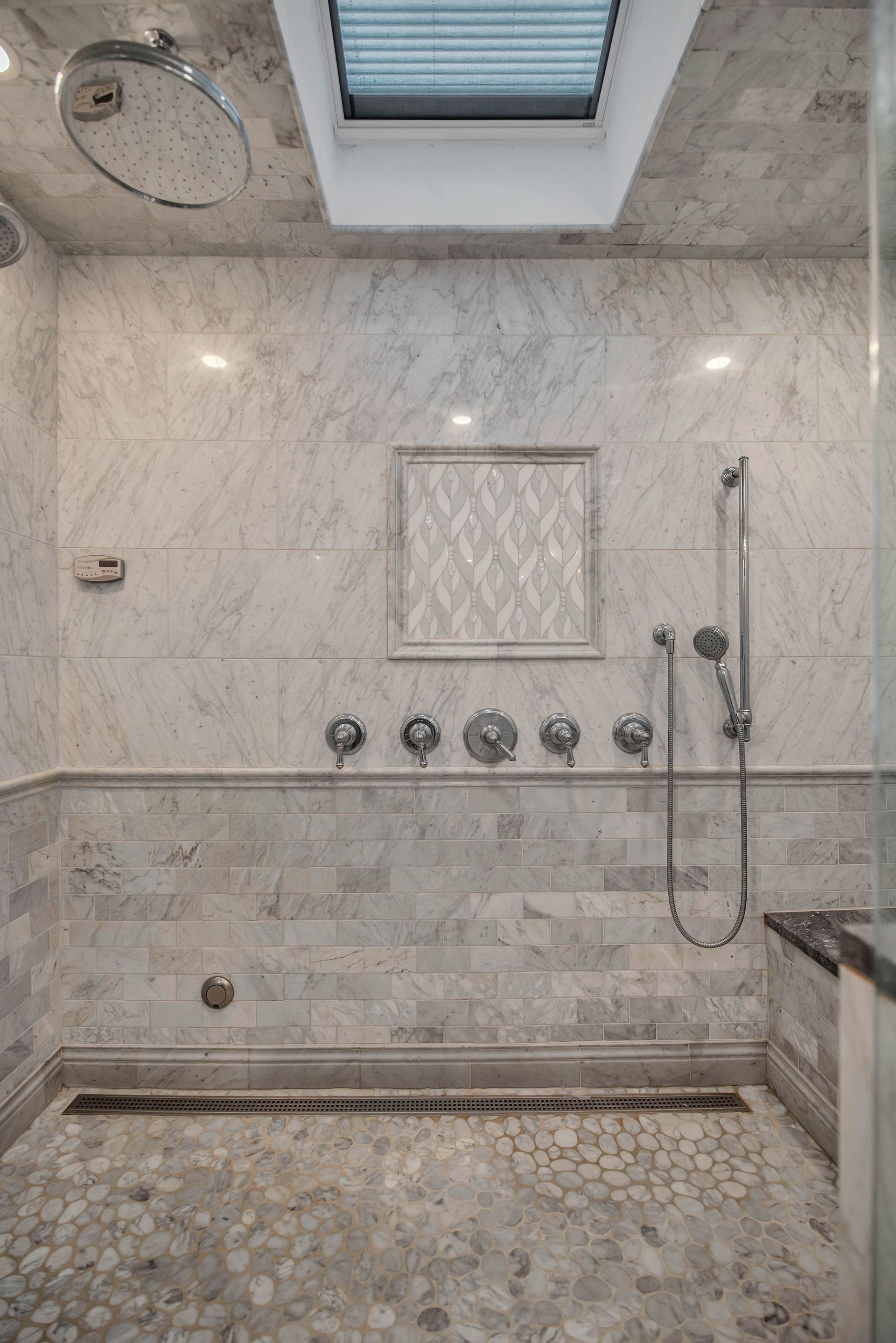 Spacious marble shower with multiple showerheads, a tiled niche, and a pebble floor. Skylight above.