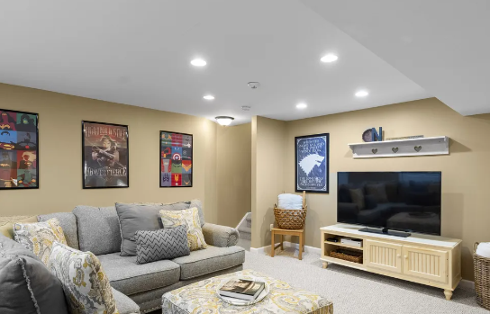 Cozy living room with gray sofa, TV, and decorative posters on light tan walls; recessed lighting.
