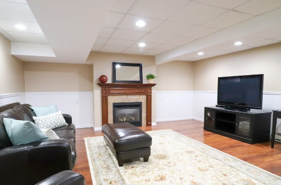 Cozy basement living room with fireplace, TV, leather sofa, and patterned rug. Warm neutral colors.