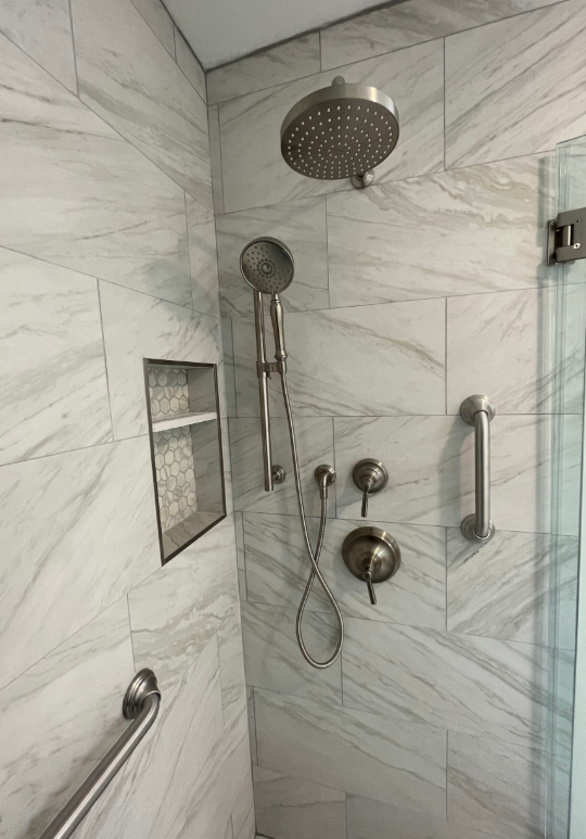 Modern shower with marble-like tile, overhead and handheld shower heads, grab bars, and a built-in shelf.