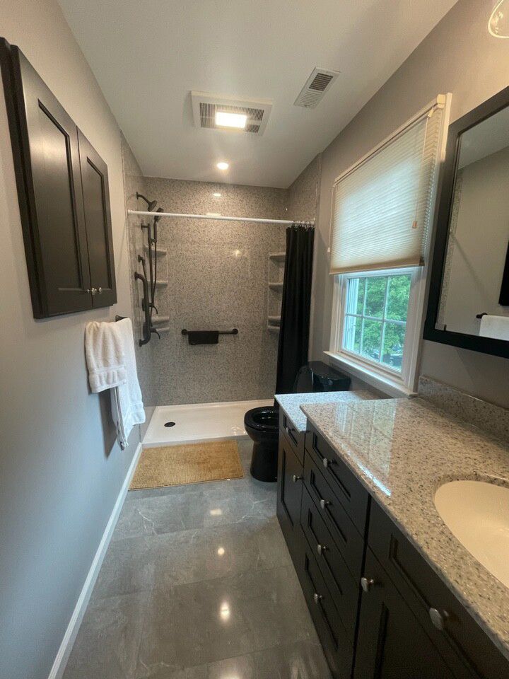 Long, narrow bathroom with gray tile floor and walls; dark wood cabinets and black toilet; shower with gray tile, open shelving.