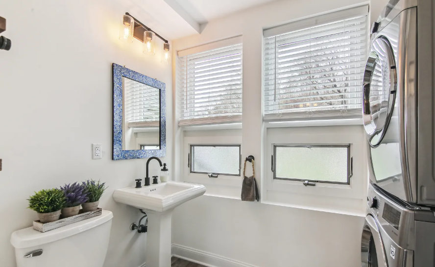 Small white bathroom with a sink, mirror, windows, washer/dryer, and a toilet with faux plants.