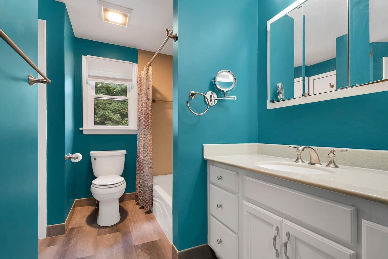 Teal bathroom with white vanity, toilet, and window; brown wood-look flooring and shower curtain.