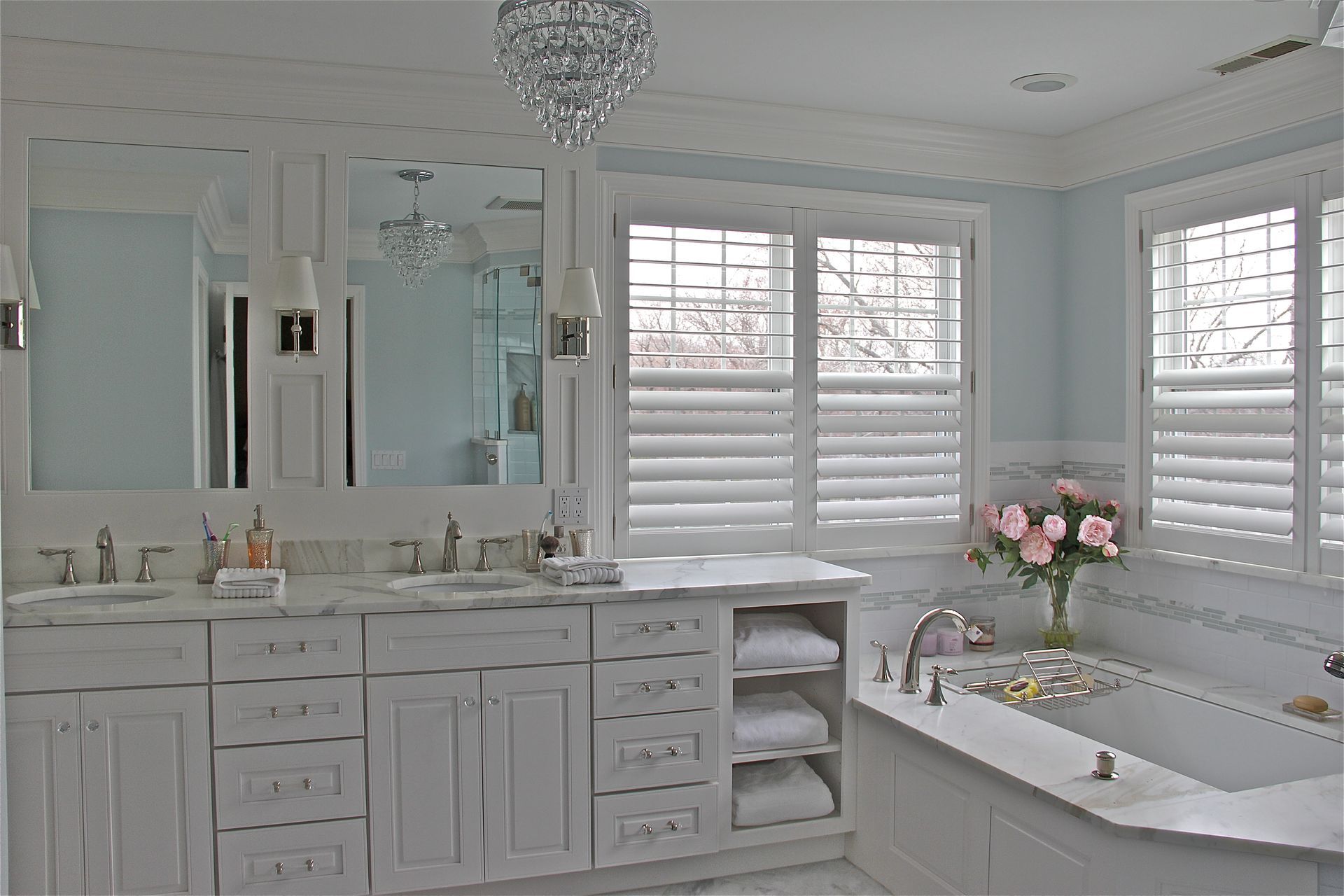 Elegant white bathroom with double vanity, bathtub, and window shutters. Features a chandelier and light blue walls.