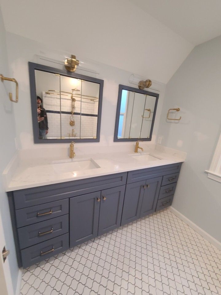 Blue double vanity with gold fixtures, white countertop, mirrors, and patterned floor in a bathroom.