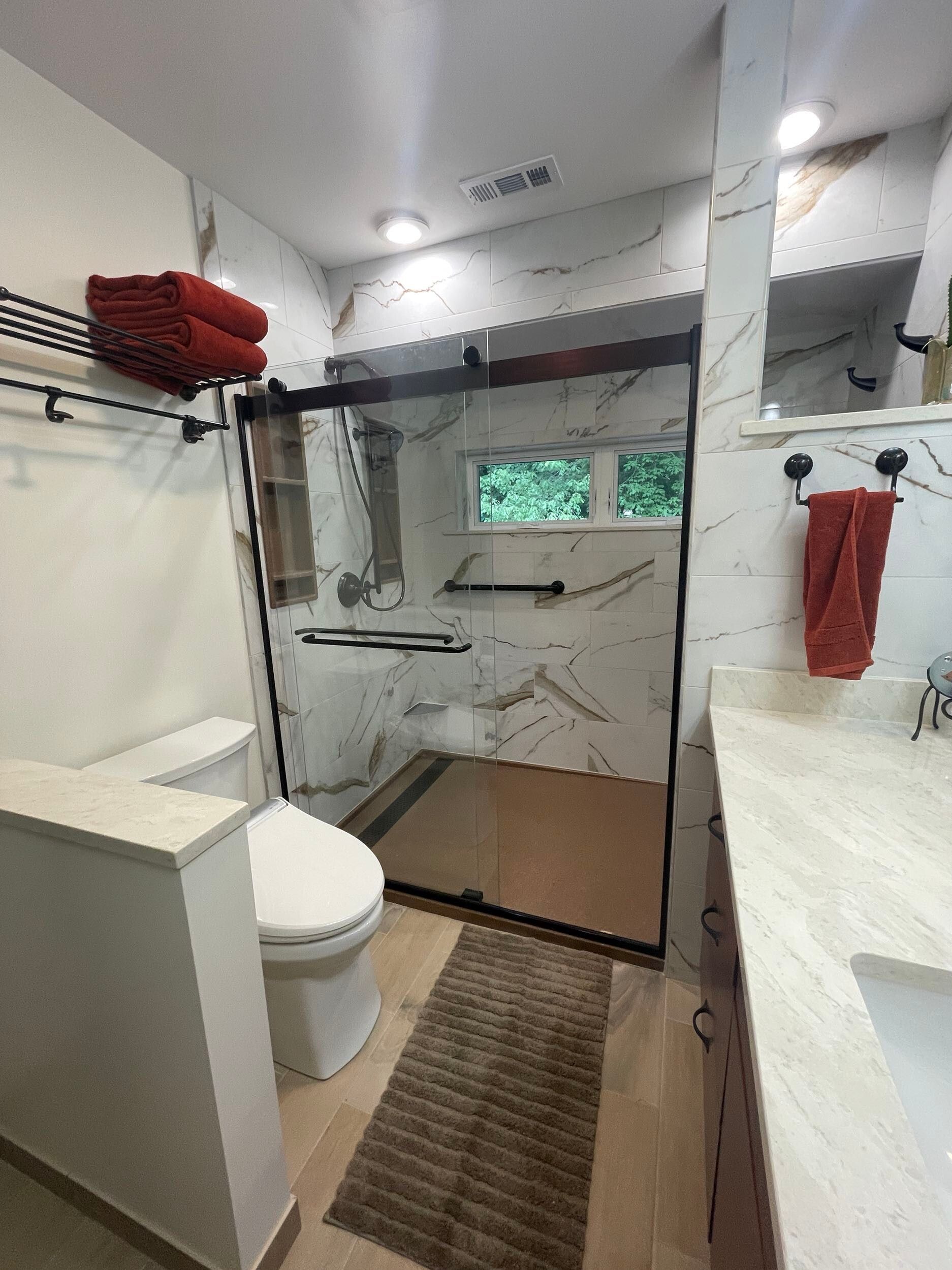 Modern bathroom with marble walls, a black-framed shower, and a toilet. Red towels add color.