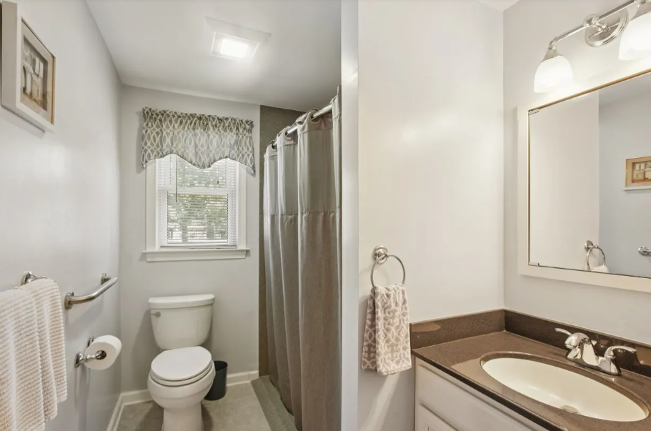 Bathroom with light gray walls, toilet, vanity, shower curtain, and window with patterned valance.