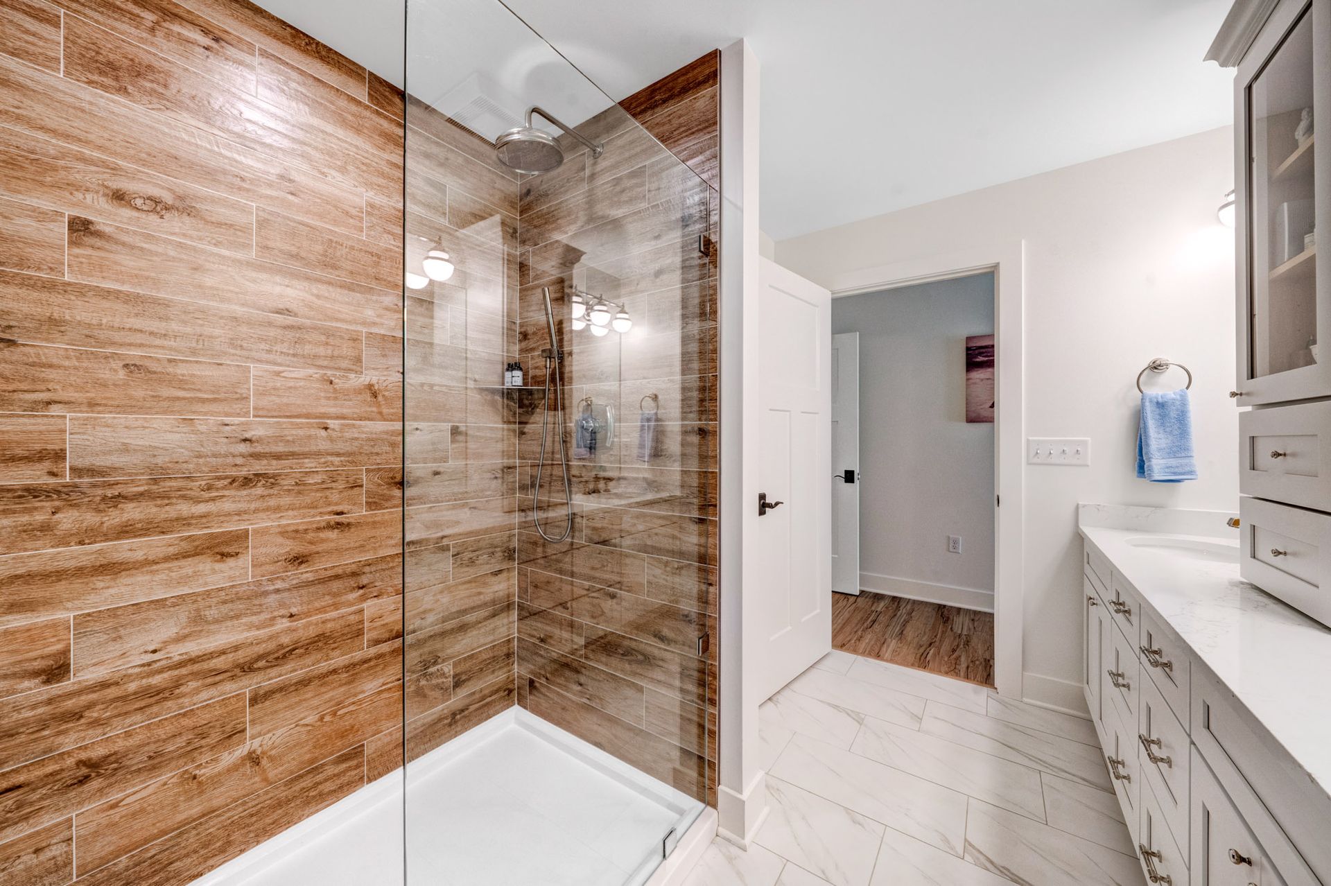 Bathroom with wood-look tile shower, glass door, white vanity, and open door to the hall.